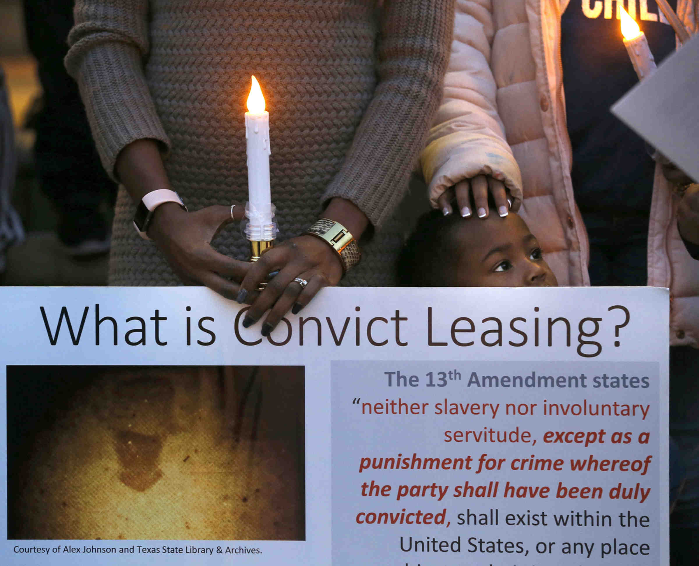 Andrea Collins, with her children, holding a sign explaining convict-leasing at a candlelight vigil, in Sugar Land, Texas on Dec. 16, 2018, honoring the 95 African-American remains found at the Fort Bend ISD construction site (Associated Press)
