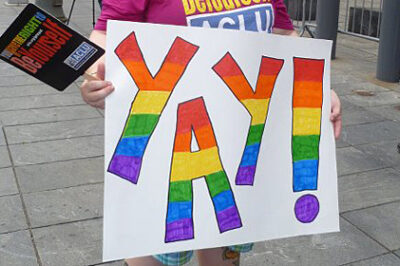 A person holding a sign reading "Yay!" in rainbow letters.