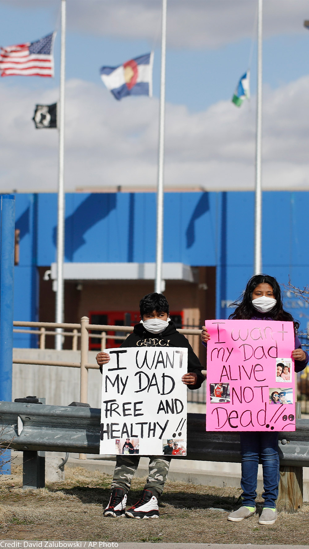 Two children wear masks and hold signs that read "I want my dad free and healthy" and "I want my dad alive not dead!!" as part of a protest calling for the release of detained immigrants in front of the GEO Detention Center in Aurora, Colorado.