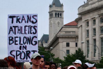A crowd of marchers with one holding a sign with the text " Trans Athletes Belong in Sport."