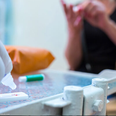 A gloved hand adds reagent to a COVID-19 serological antibody test with the patient sitting nearby.