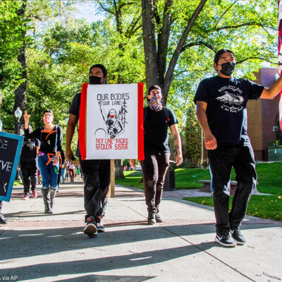 A group of people marching holding signs and a flag. The signs have text that read "You're on native land #landback" and "Our bodies our land / Not one more stolen sister".