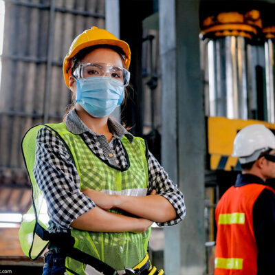 A technician wearing a surgical mask and hard hat stands with arms crossed in a factory.