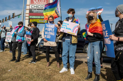 Protestors holding an ACLU "Trans People Belong" poster and "trans kids are kids" poster at a trans rights rally in South Dakota.