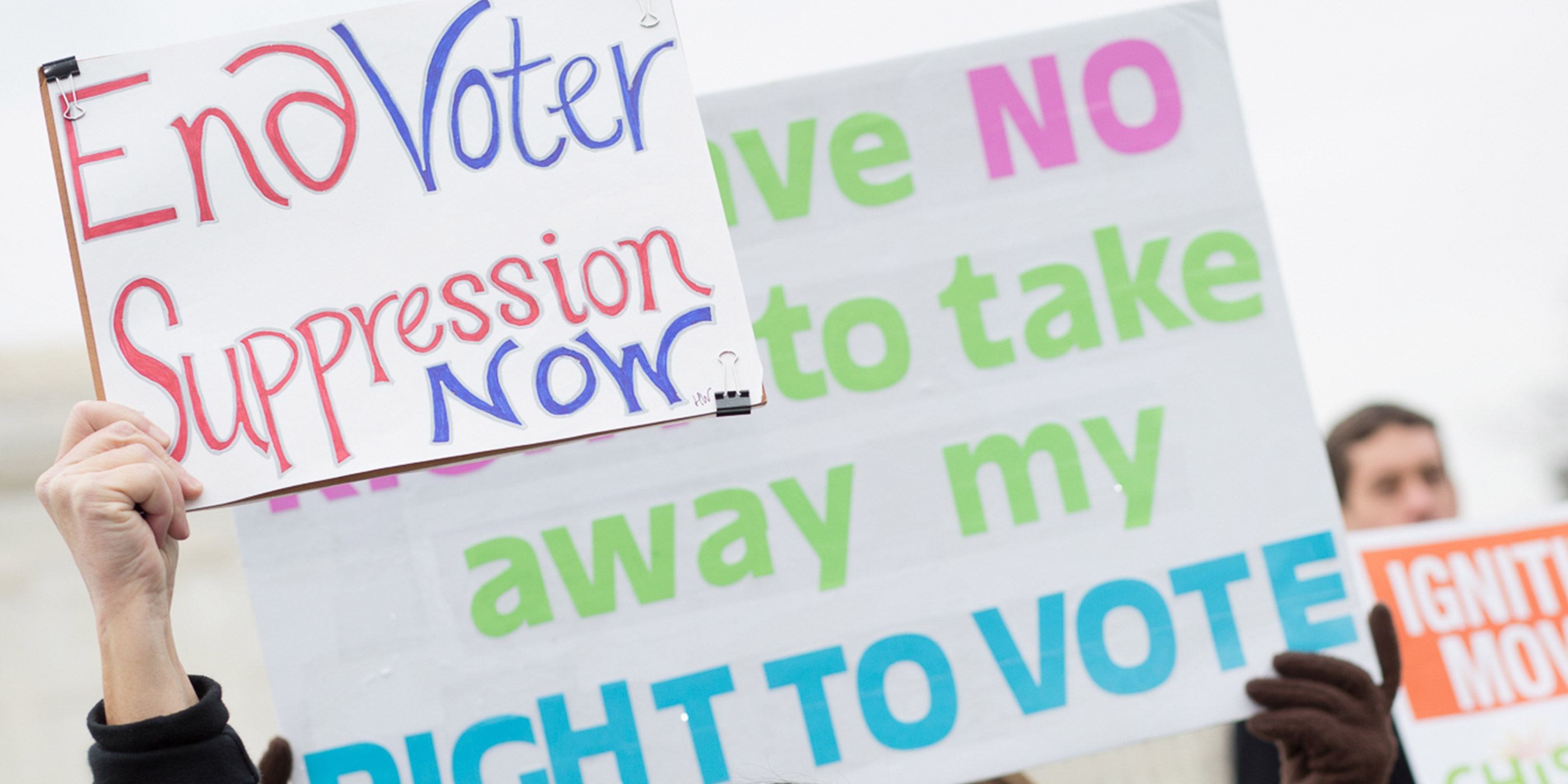 Demonstrators hold signs, one of which says “End Voter Suppression Now.”