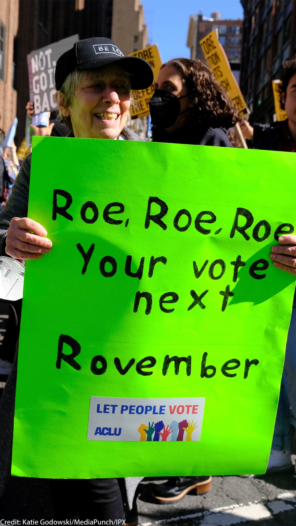 A woman holding a sign reading "Roe, Roe, Roe, Your vote next Rovember / LET PEOPLE VOTE ACLU" poses for the camera as fellow activists walk behind her.