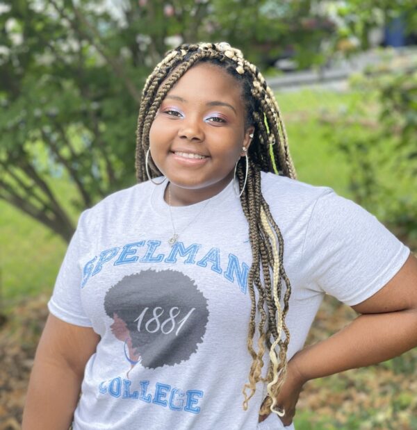 Amara Harris smiling, standing in front of a tree. She is wearing a Spelman College t-shirt and hoop earrings.