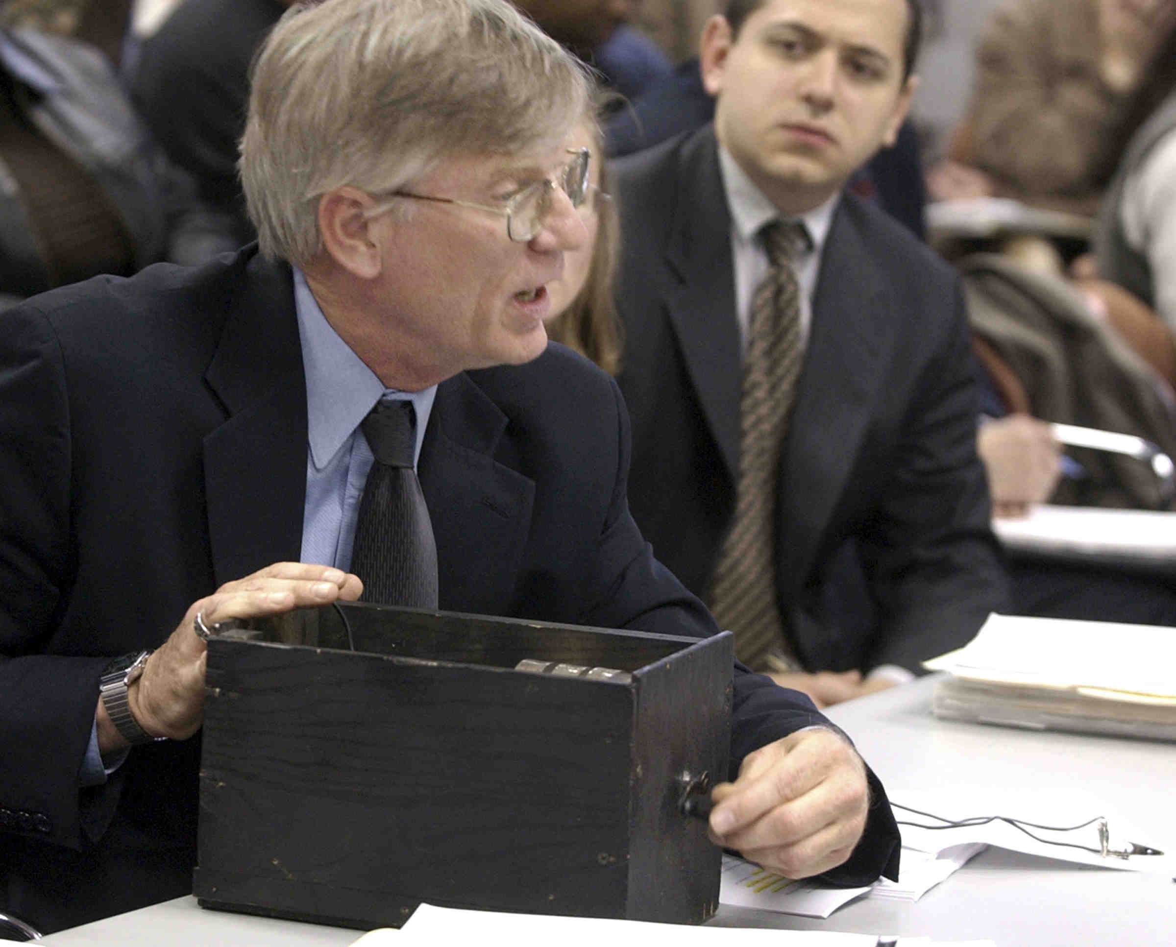 A man in a suit speaking while holding a black box