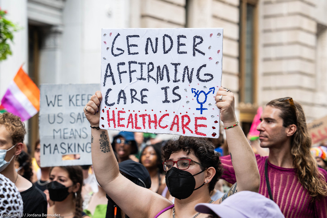 A shirtless demonstrator (wearing glasses and a black surgical mask) holds up a sign that reads" GENDER AFFIRMING CARE IS HEALTHCARE".