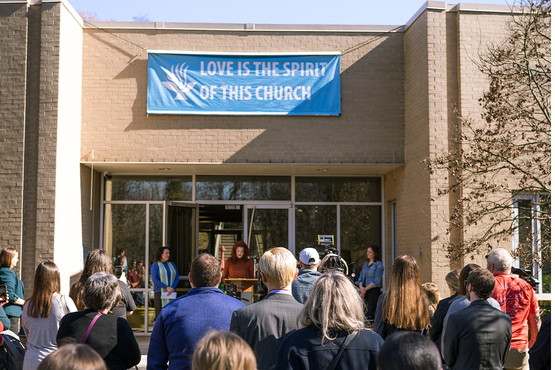 People (with their backs to the camera) walk towards a nondescript building's glass doors. where above the doors hangs a sign that reads "LOVE IS THE SPIRIT OF THIS CHURCH".