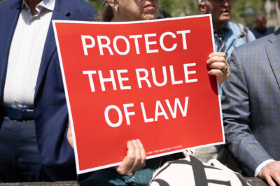 At the Lawyers Rally In Lower Manhattan To Demand Respect For The Rule Of Law, a demonstrator holds up a sign with white lettering on a red background that reds, "PROTECT THE RULE OF LAW".