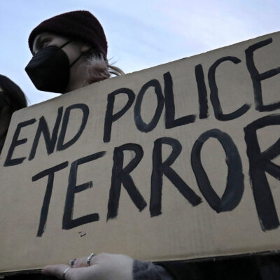 A demonstrator wearing a black hospital mask and black beanie hat holds up a cardboard sign that reads, "END POLICE TERROR".