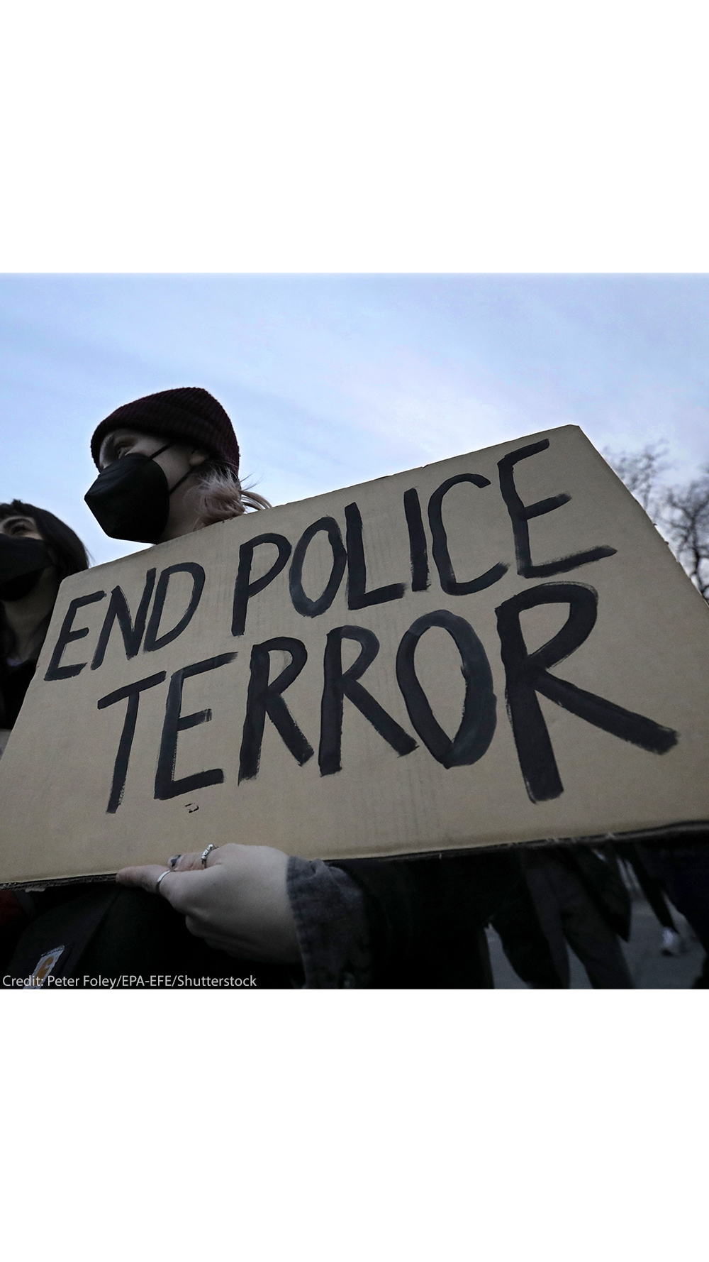 A demonstrator wearing a black hospital mask and black beanie hat holds up a cardboard sign that reads, "END POLICE TERROR".