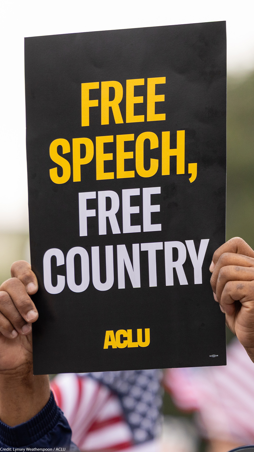 A demonstrator (with only their hands visible) holds up a sign that reads " FREE SPEECH, FREE COUNTRY - ACLU".