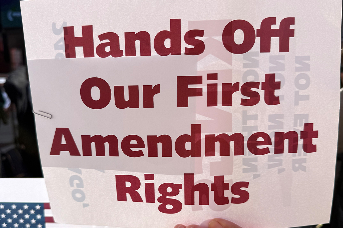 A demonstrator (with only their hand visible) holds up a sign that reads "Hands Off Our First Amendment Rights".