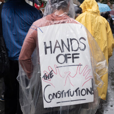 A person wearing a clear poncho displays a sign on their back that reads "HANDS OFF THE CONSTITUTION" while walking in the rain as they take part with thousands of other New Yorkers take part in the NO KINGS national day of action.
