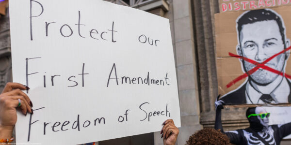 A person holds a large handwritten sign reading “Protect our First Amendment Freedom of Speech” during a protest outside the El Capitan Entertainment Centre, where Jimmy Kimmel Live! is filmed. Behind them, another protester in a skeleton costume holds a cardboard sign depicting a crossed-out image of Jimmy Kimmel. The demonstration is in response to ABC’s decision to pull Kimmel off the air following his remarks about Charlie Kirk.