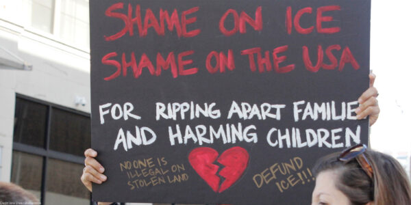 A demonstrator protests ICE and the Trump Administration policy of separating migrant families by holding up a sign (written in red ink on a black background with an illustrated broken red heart at the bottom) that reads, "SHAME ON ICE SHAME ON THE USA FOR RIPPIING APART FAMILIES AND HARMING CHILDREN No One is Illegal On Stolen Land, Defund ICE!!!"