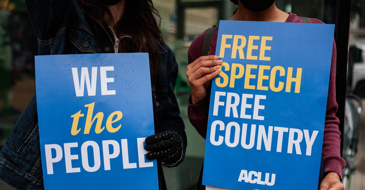 Two protesters hold signs that read Free Speech Free Country and We The People.