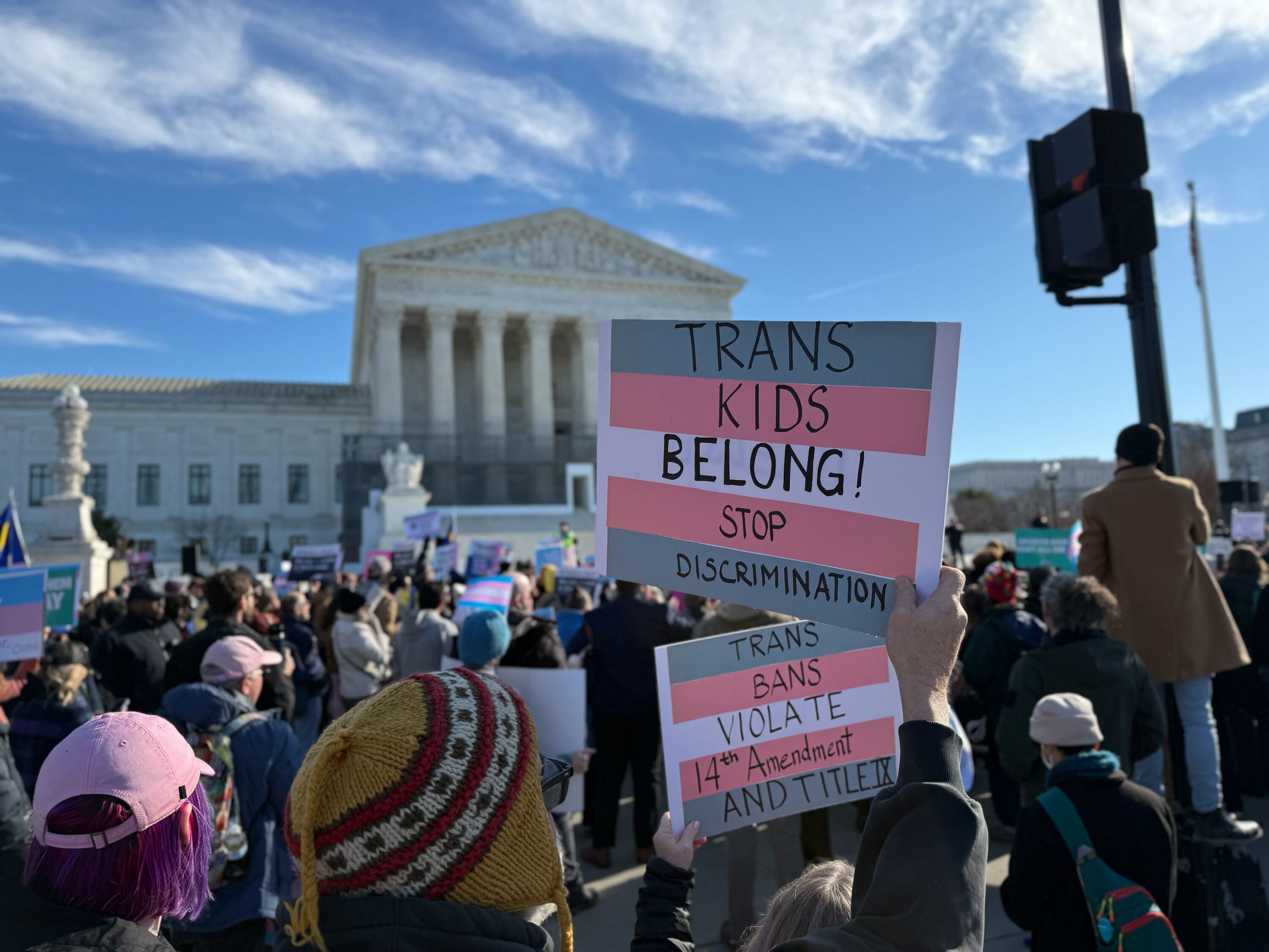 Protestors stand in front of the Supreme Court holding signs that say "Trans kids belong! Stop Discrimination!" and "Trans Bans Violate 14th Amendment and Title IX" printed on the trans pride flag.