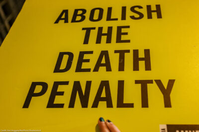 Yellow protest sign with bold black text reading “Abolish the Death Penalty,” partially held by a person at the bottom edge.