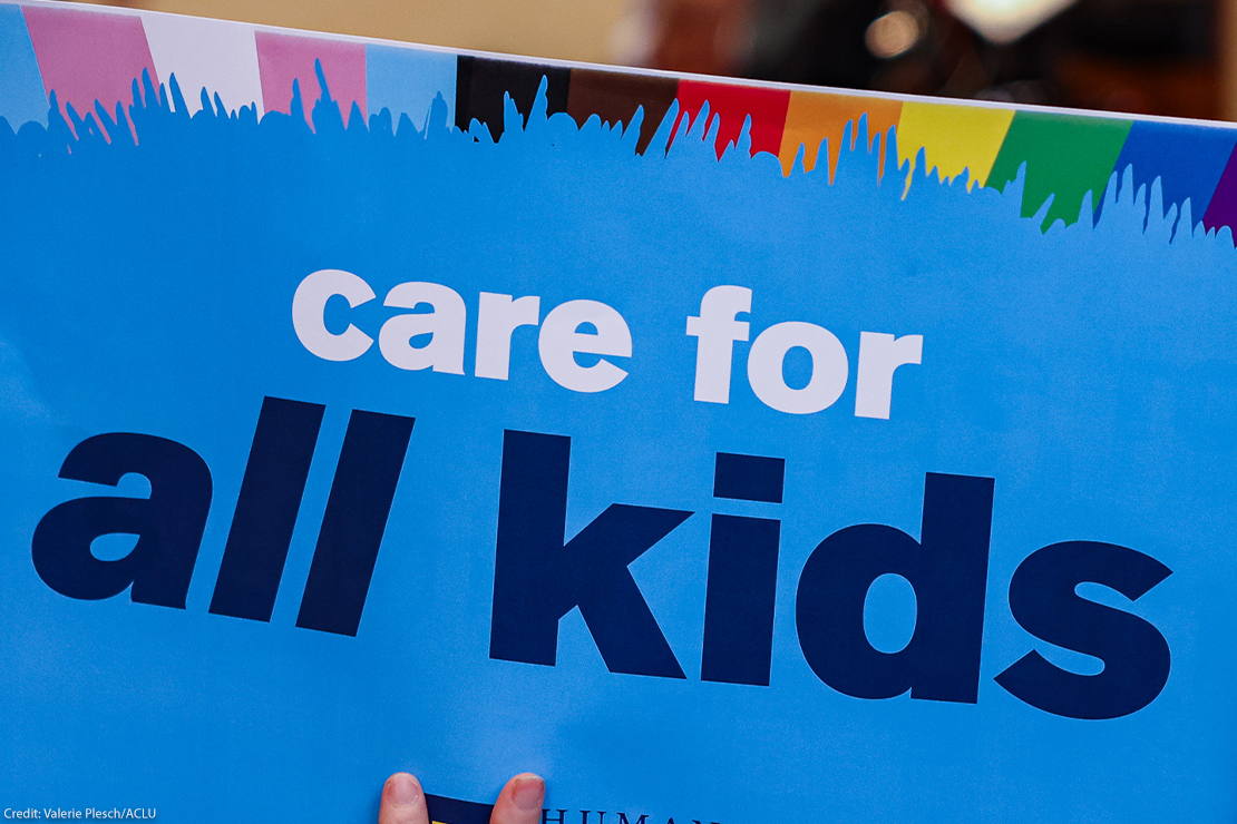 Close-up of a blue protest sign reading “care for all kids,” with a rainbow-colored border along the top edge and hands holding it at the bottom.